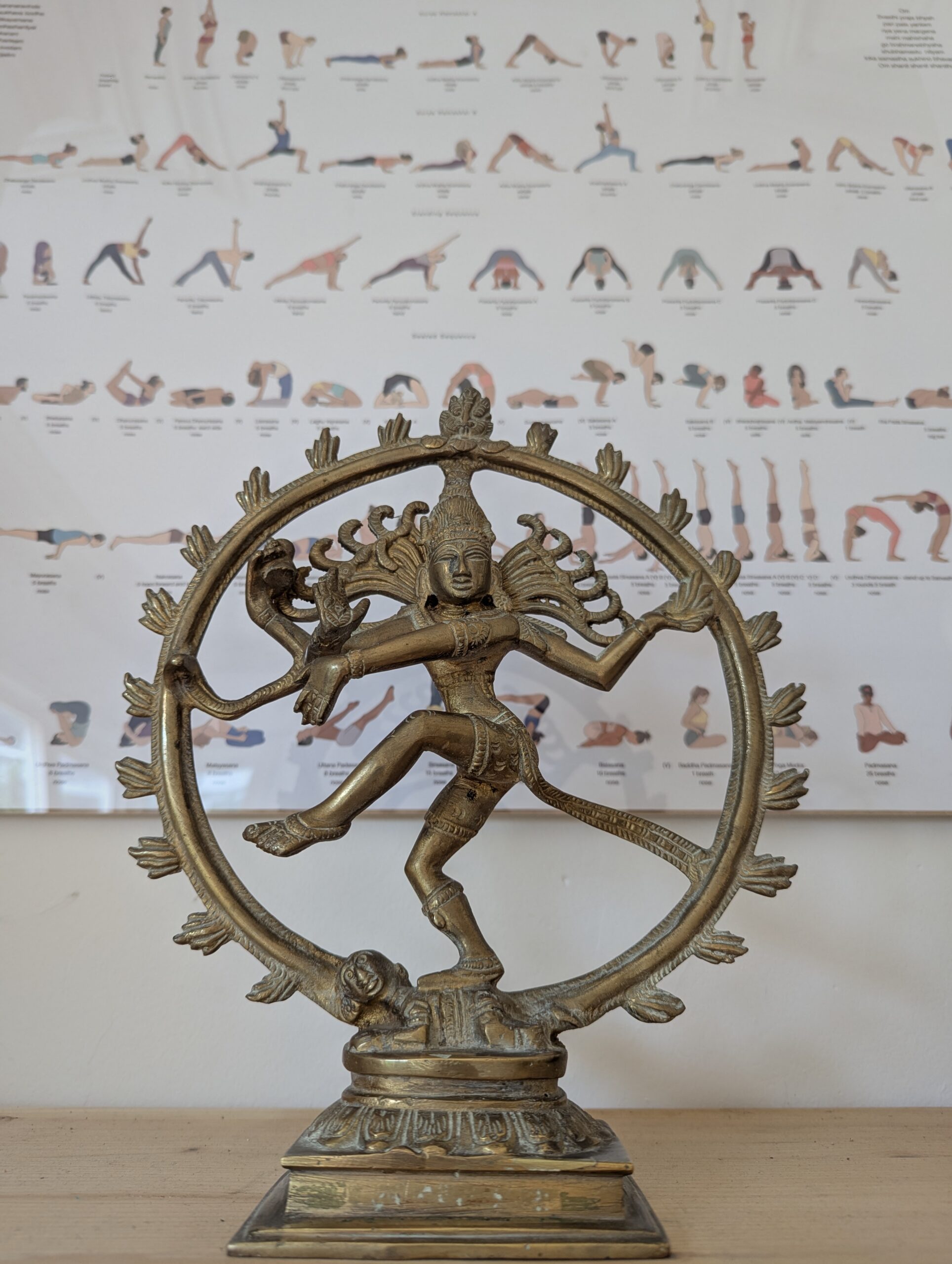A photograph captures an intricately detailed brass statue of the Hindu deity Shiva in the form of Nataraja, the Lord of the Dance. The statue is positioned centrally on a light-coloured wooden shelf, showing Shiva in a dynamic cosmic dance pose with multiple arms (four clearly visible) within a large circular, flame-fringed halo. He balances on his right leg on a prostrate figure, and his left leg is raised. The background, which is slightly out of focus, features a framed chart pinned to a white wall, displaying rows and rows of small illustrative human figures in various yoga asanas (poses), arranged in a sequence with unreadable descriptive text below each pose. The statue has a worn, aged metal finish. The perspective is frontal.